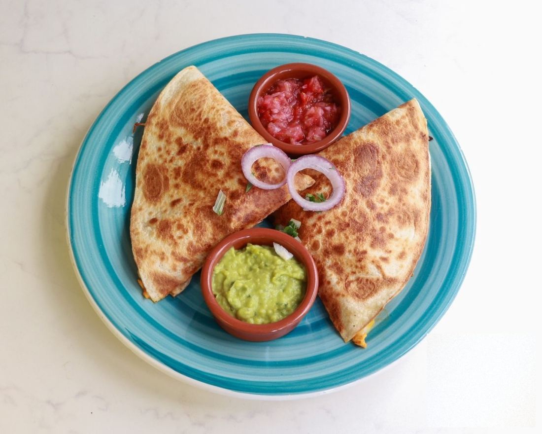Deux quesadillas dorées coupées en deux sur une assiette turquoise, avec guacamole, pico de gallo et oignon rouge.