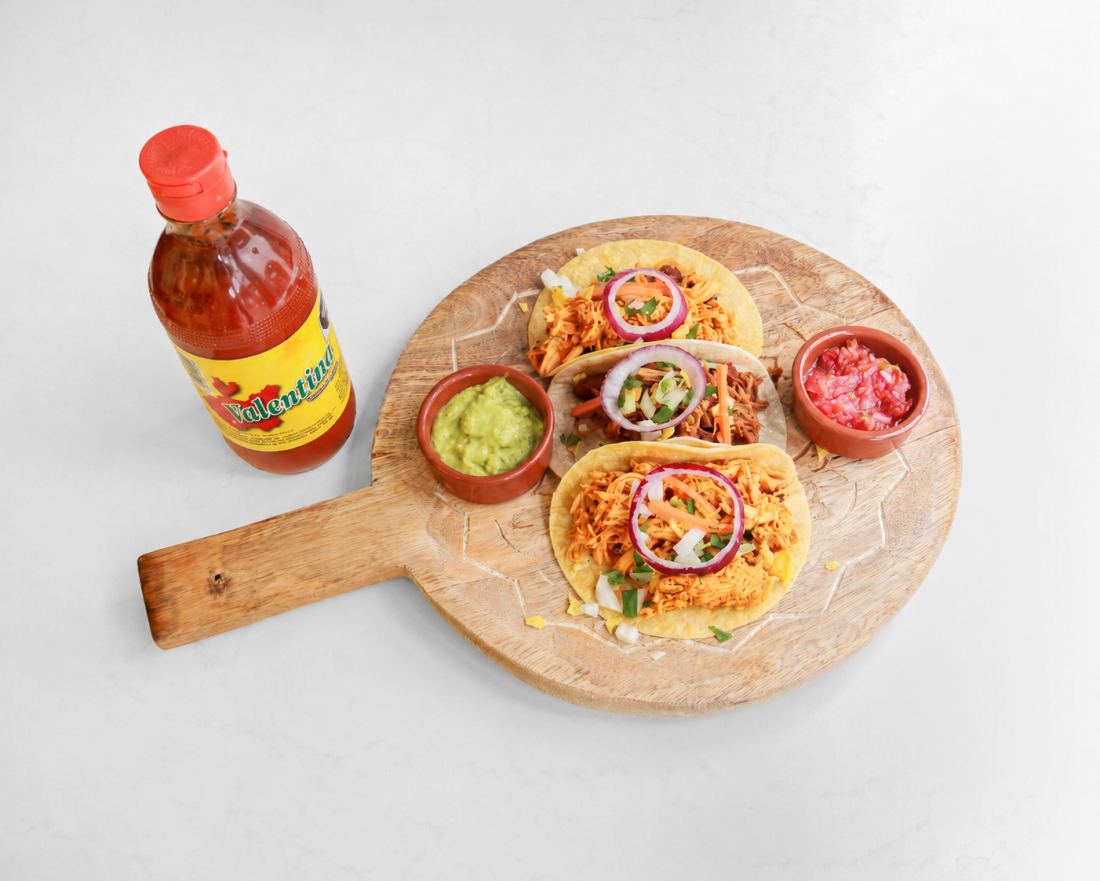 Trois tacos mexicains sur une planche en bois, garnis de viande effilochée, oignon rouge et coriandre, avec guacamole, pico de gallo et sauce piquante.