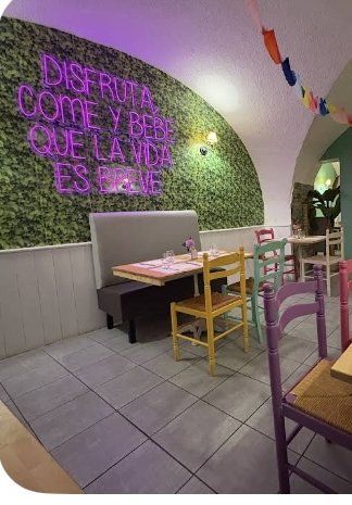 Un coin de la salle du Barrio Latino : banquette, tables dressées, chaises dépareillées jaune et violette devant le mur végétal et le néon rose.