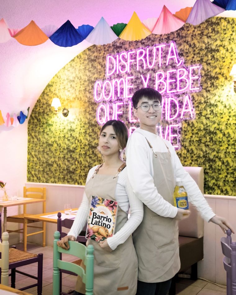 Gianella et Clément Pélissier, dos à dos dans la salle du Barrio Latino, devant le néon rose « Disfruta, come y bebe que la vida es breve » et le mur végétal, sous une guirlande d’éventails en papier multicolores.