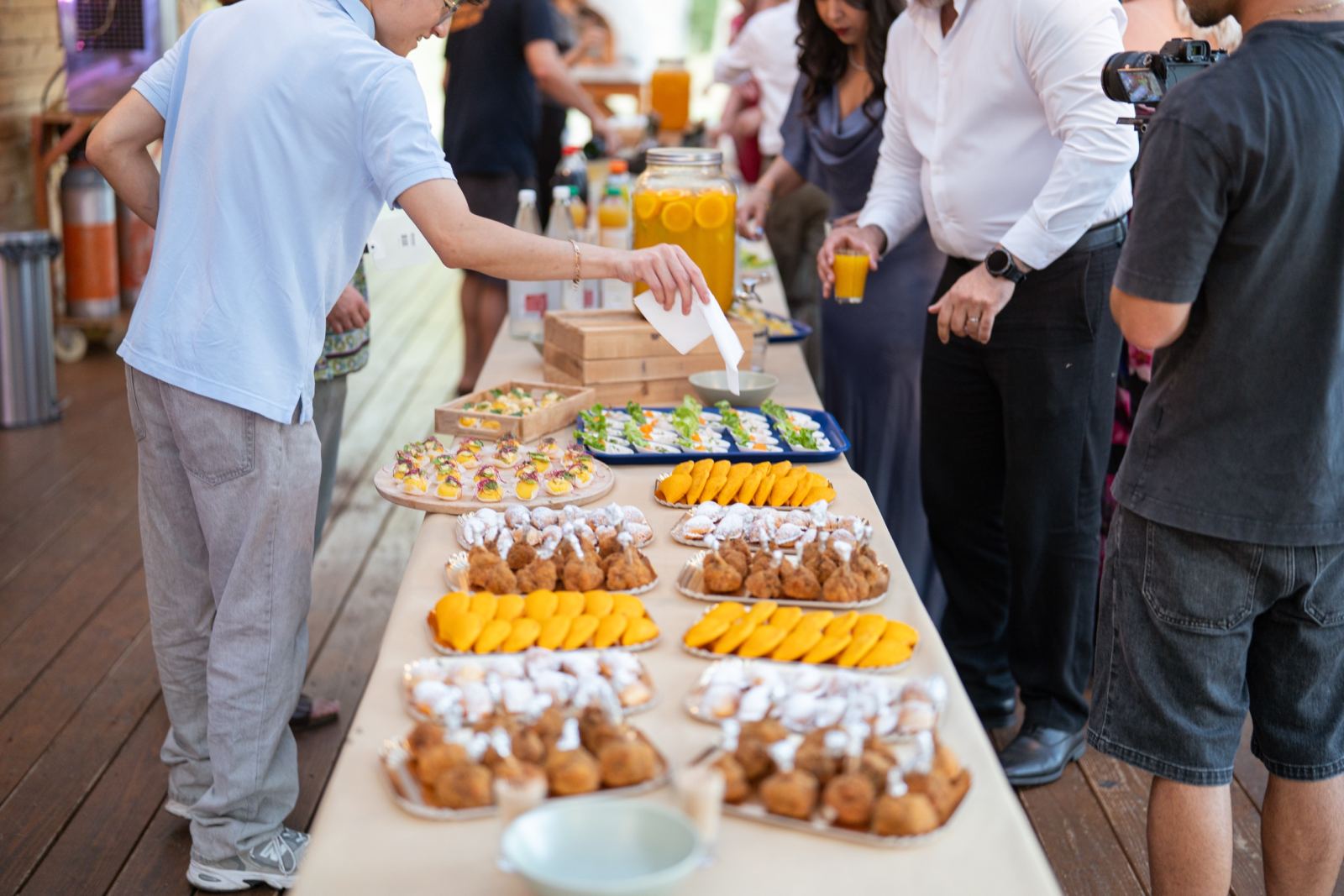Un long buffet dressé sous une terrasse en bois lors d’un événement : plateaux d’empanadas, verrines de ceviche et grande bonbonne de jus d’orange, les invités se servent.